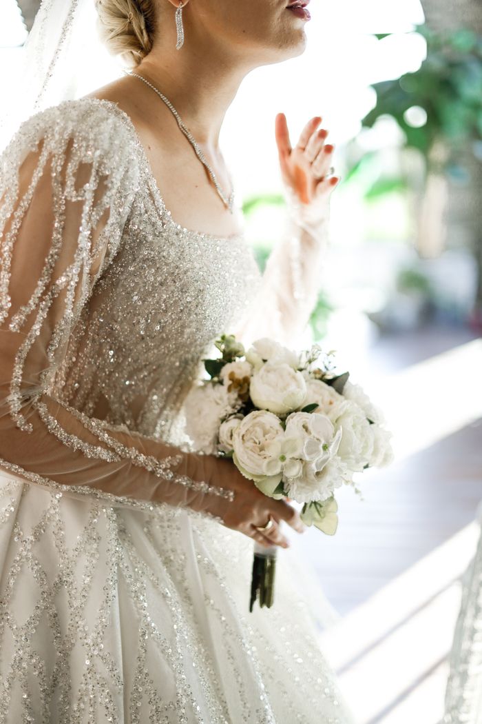 A bride in a glittering gown holding a white bouquet in a sunlit room.
